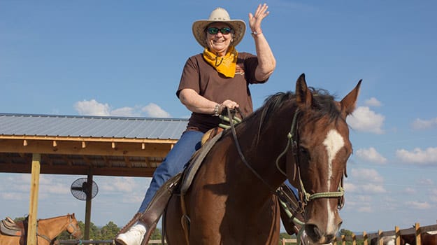 Senior riding a horse outdoors, waving happily.