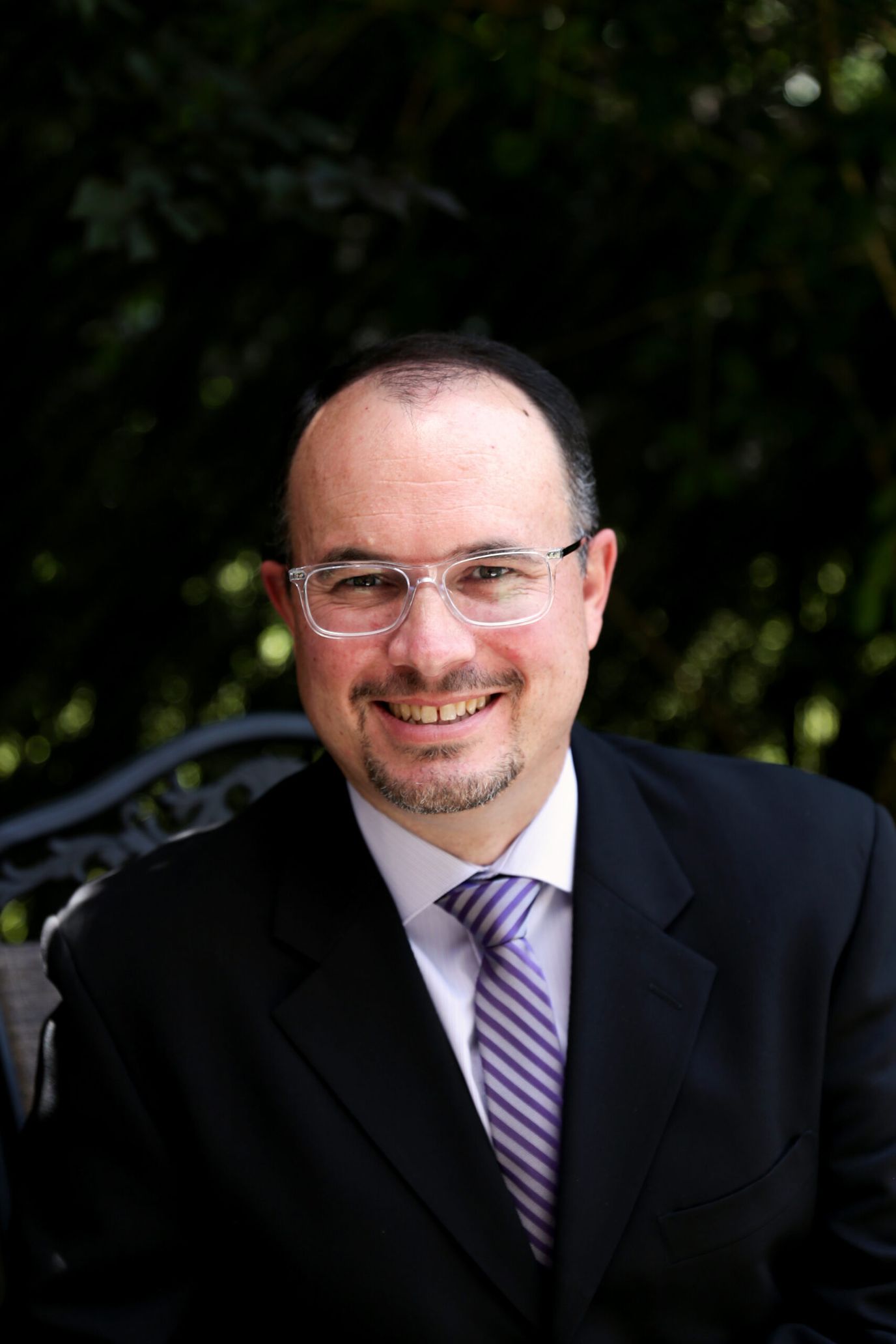 A man in a suit with glasses smiling outdoors