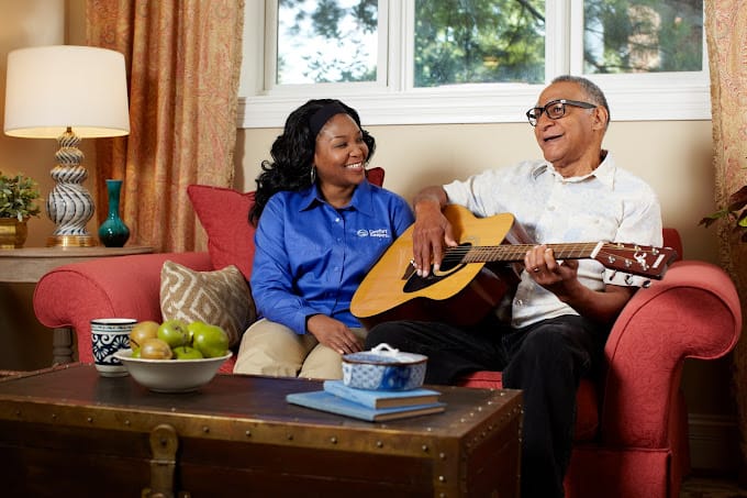 A caregiver and a senior man enjoying music together while sitting on a couch.