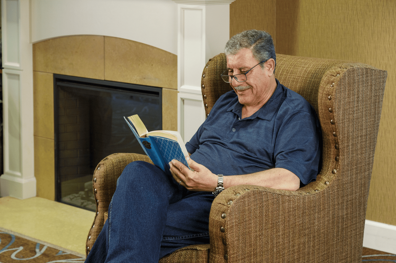A resident reading a book in a cozy chair by the fireplace