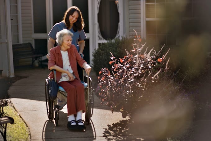 A caregiver pushing a resident in a wheelchair along a garden path.