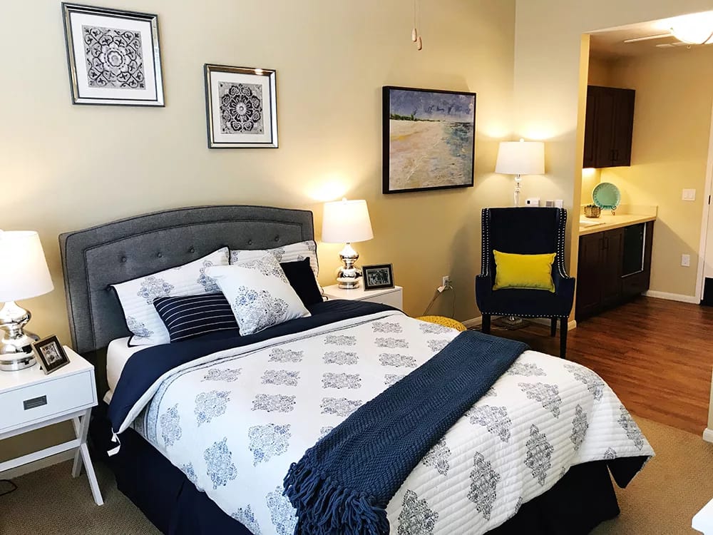 A well-decorated bedroom featuring a gray upholstered bed with striped and patterned pillows, bedside lamps, and a cozy chair.