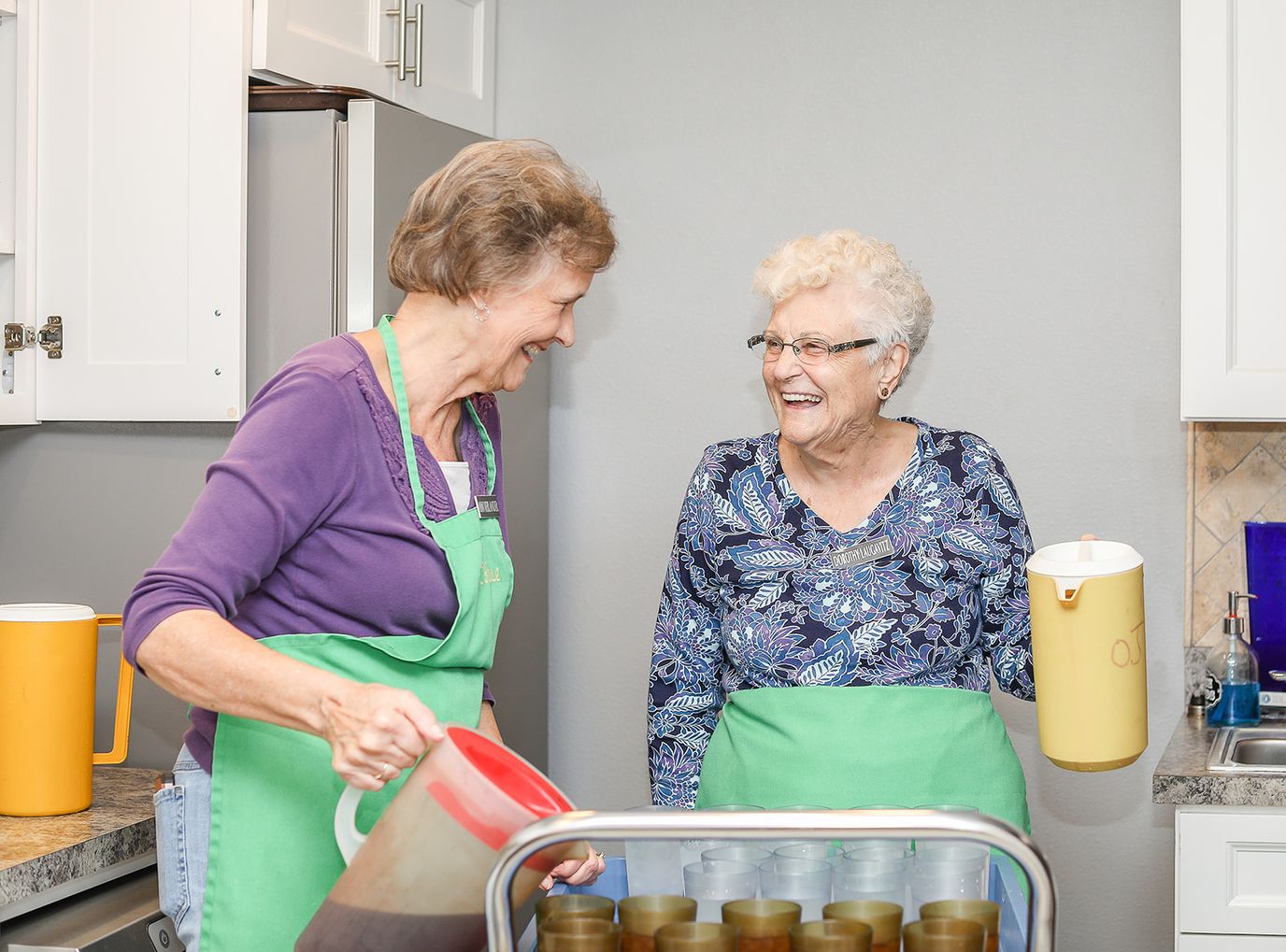 Two staff members smiling and pouring drinks in a kitchen