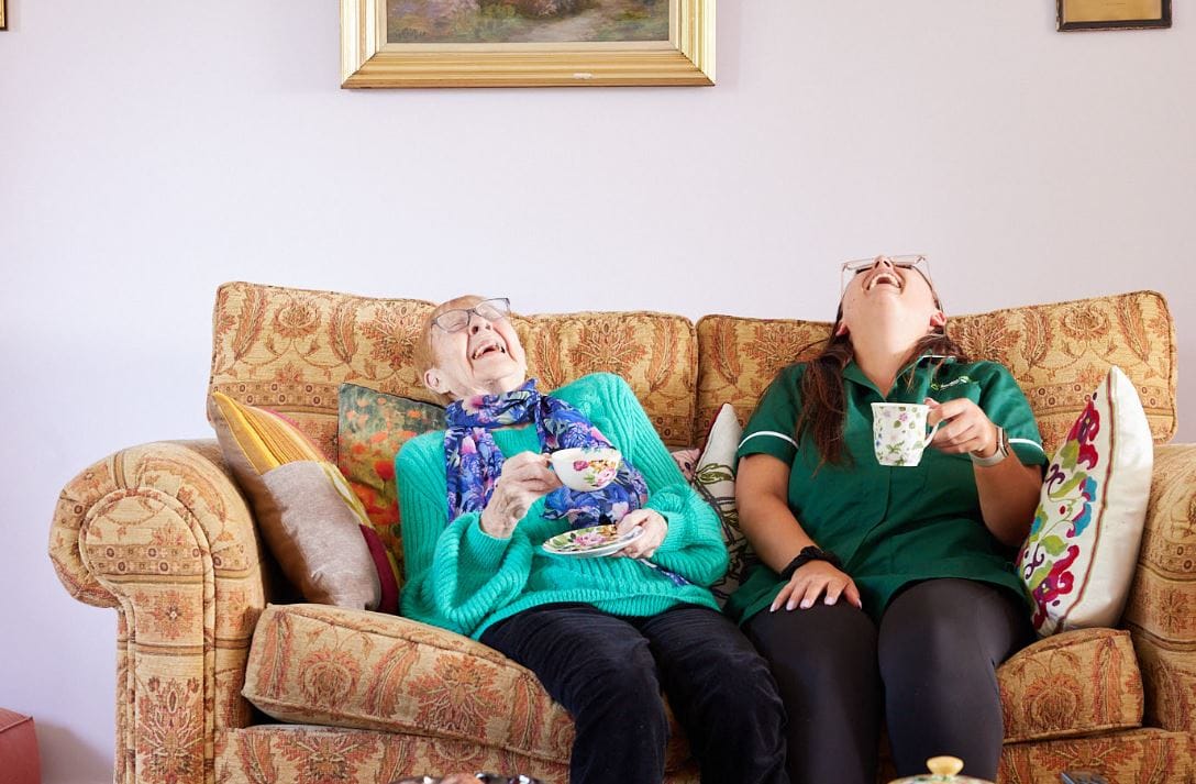 Resident and staff laughing together on a cozy couch