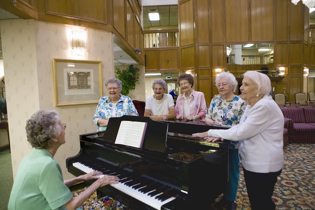 Six senior women gathered around a piano with one playing