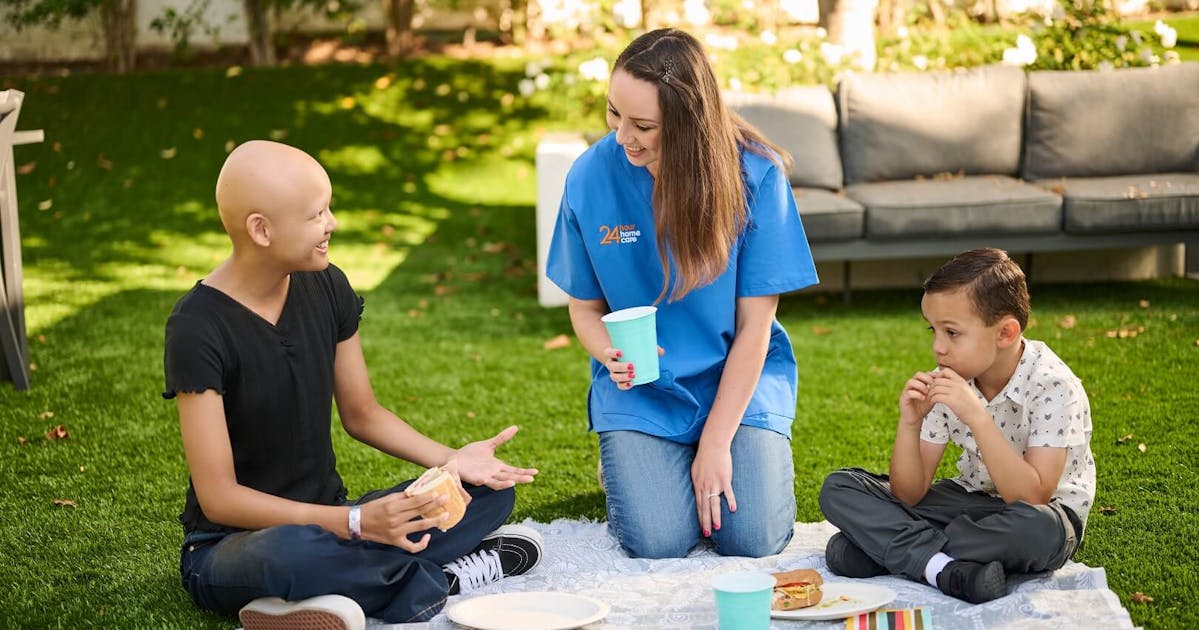 A caregiver and two children enjoying snacks outdoors.