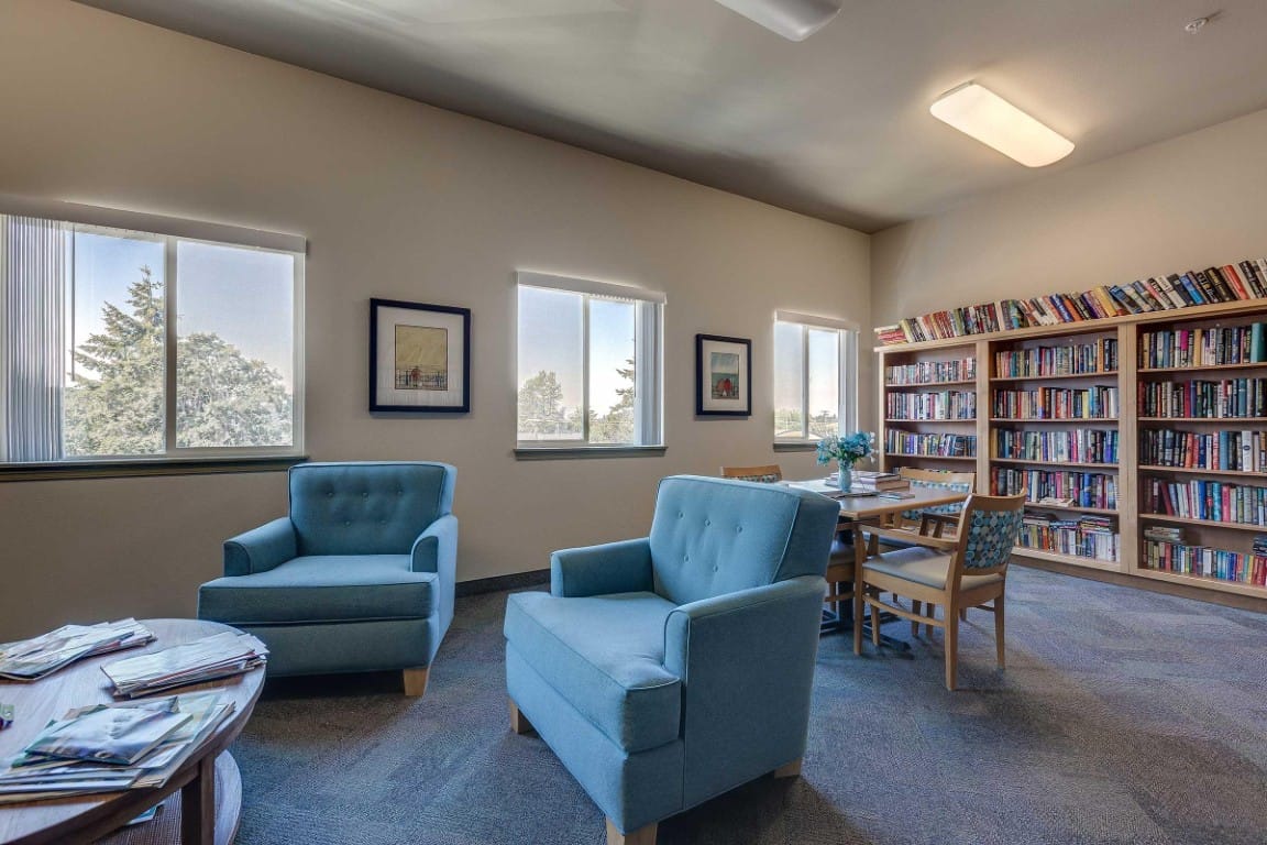 A bright library lounge with blue chairs, a wooden table, and bookshelves filled with books.