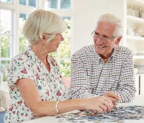 Two seniors smiling and doing a puzzle indoors