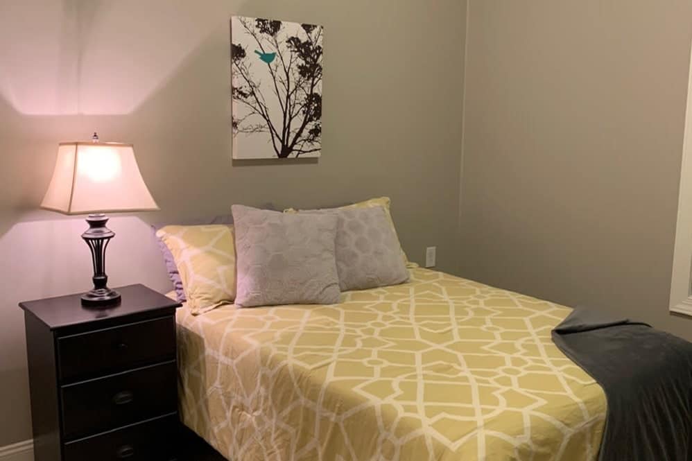 A well-lit bedroom featuring a bed with yellow geometric patterned bedding and a bedside table with a lamp.