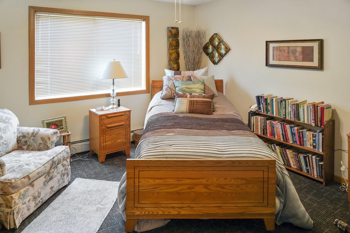 A cozy bedroom featuring a bed with pillows, a side table with a lamp, and a bookshelf.