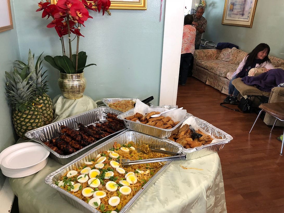A table with various food trays, including fried chicken, rice, and garnished eggs, with a pineapple and flowers in the background.