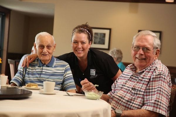 Two senior men smiling with a staff member at a dining table.