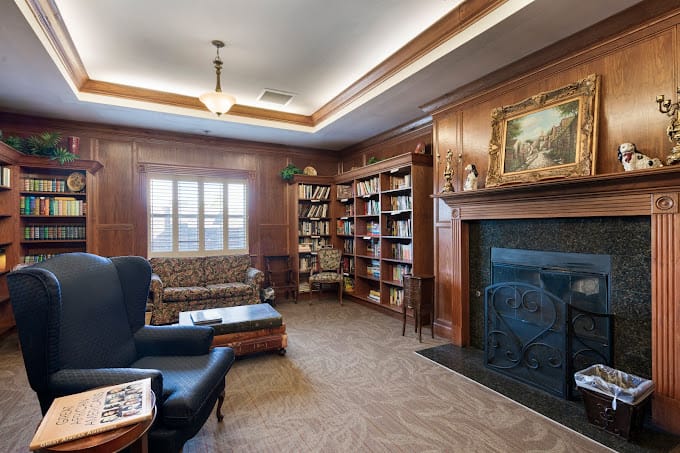 A cozy library with bookshelves, a fireplace, and seating at Heritage Manor Stratmore.