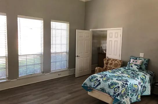 Brightly lit bedroom with a bed and chair, large windows.