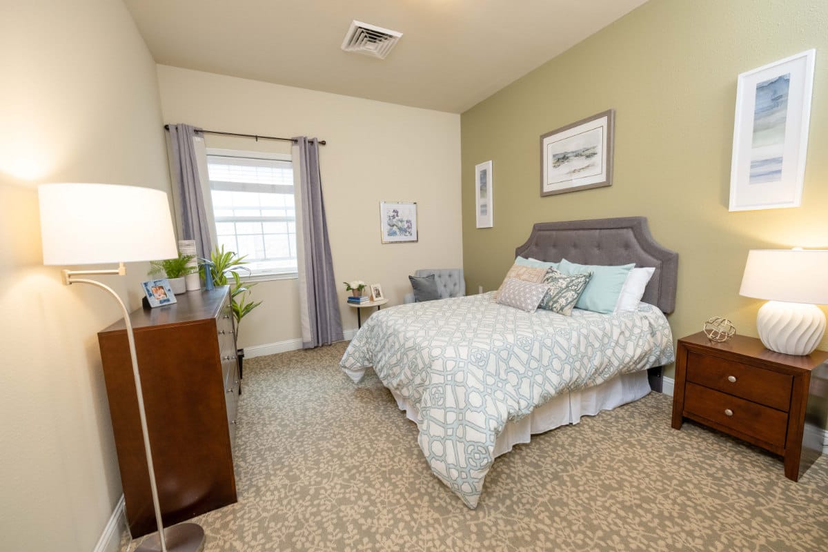 A well-decorated bedroom with a bed, dresser, and lamp in The Heritage Memory Care.