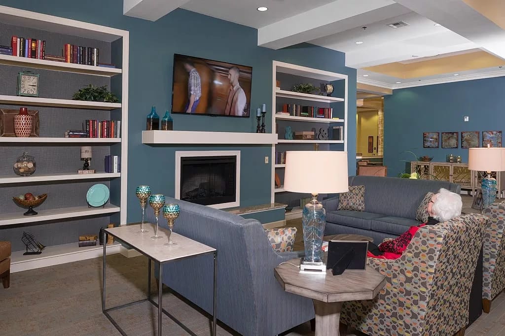 A senior woman relaxing in a stylish lounge area with a TV, bookshelves, and comfortable seating.