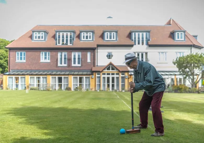 An elderly man playing croquet on the lawn in front of the building.