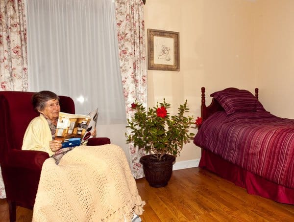 An elderly woman reading a magazine in a cozy seating area with a plant and bed nearby.