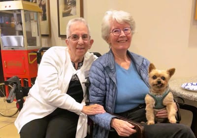 Two women smiling with a small dog in a common area.