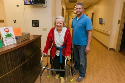 A resident using a walker with staff assistance in a hallway