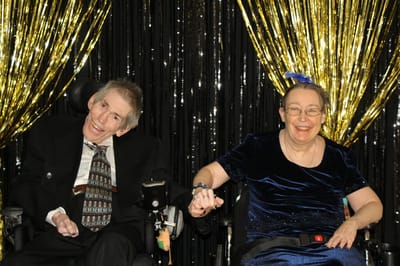 Two elderly residents in formal attire smiling and holding hands.