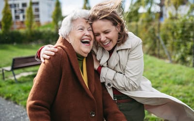 A senior woman joyfully laughing with a caregiver outdoors.