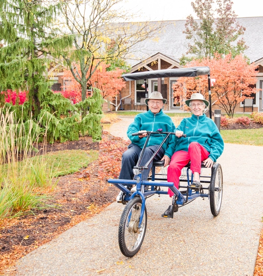 Two seniors enjoying a ride on a tricycle in the garden.