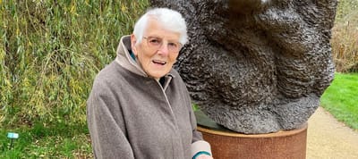 An elderly woman smiling near a sculpture in a garden