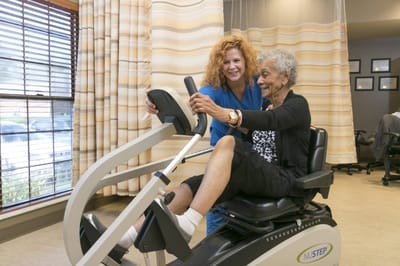 Staff assisting a resident with exercise equipment in a therapy room