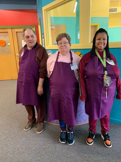 Three staff members wearing purple aprons in a colorful common area.