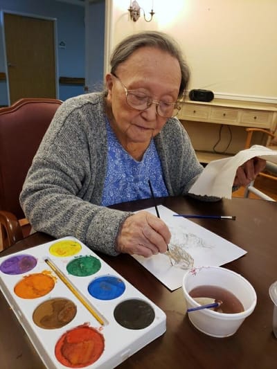 An elderly woman painting at a table with art supplies