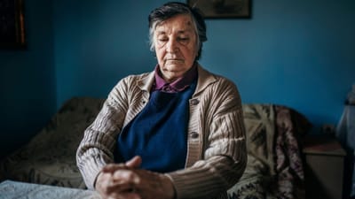 An elderly woman seated quietly indoors, reflecting.