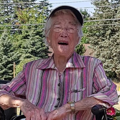 An elderly woman smiling outside with trees in the background