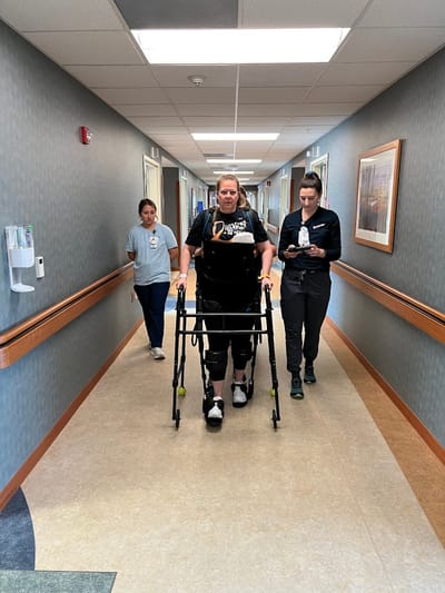 Patient using a walker assisted by staff in a hospital hallway.