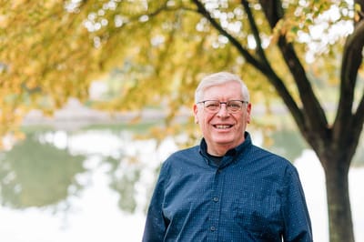 A smiling elderly man standing under a tree by a lake