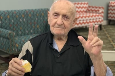 An elderly man holding a piece of cake and waving.