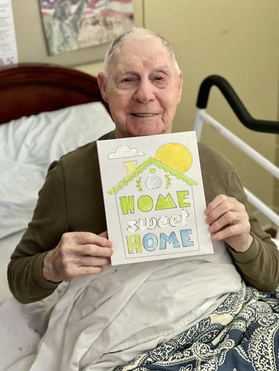 A resident holding a colorful 'Home Sweet Home' sign in bed