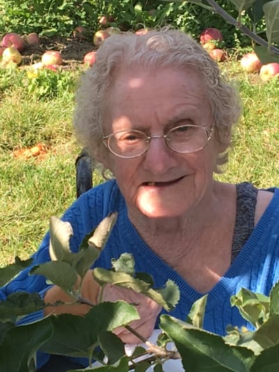 An elderly woman smiling outdoors among apple trees