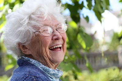 Elderly woman smiling outdoors with greenery in background