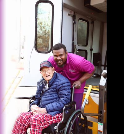 A staff member assists a resident in a wheelchair by a facility vehicle