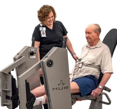 A therapy session with a senior man using exercise equipment, assisted by a staff member.