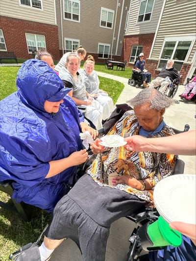 Residents enjoying food outdoors with staff assistance.