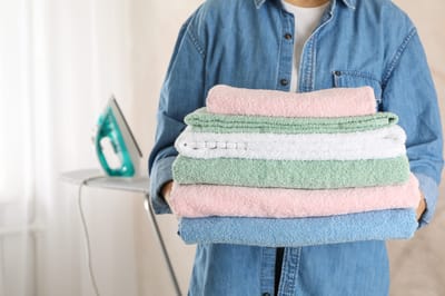 Person holding stacked towels with an iron in the background