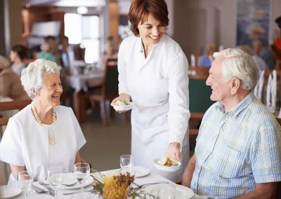 A caregiver serving a meal to smiling residents in the dining room