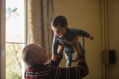 A senior man joyfully holds a baby indoors.