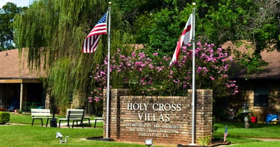 Exterior sign of Holy Cross Villas with flags and flowers.