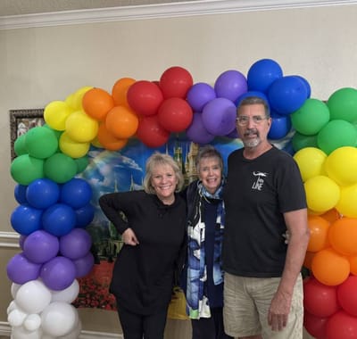 Three people smiling by a colorful balloon arch