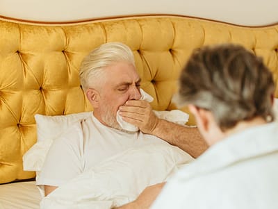 A senior man resting in bed with a caregiver nearby