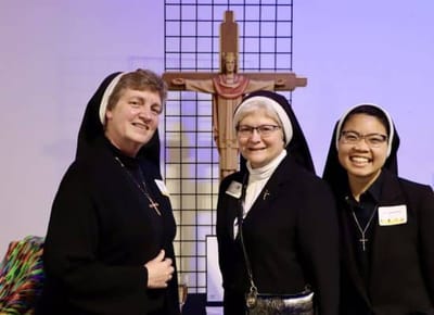 Three nuns smiling near a religious cross