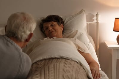 Older woman resting in bed with caregiver nearby.